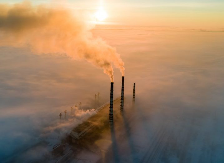 Aerial view of three pipes from a power plant blowing smoke into the air, with lots of smoke around it and the sun in the distance. Used to represent carbon and the use of fossil fuels.