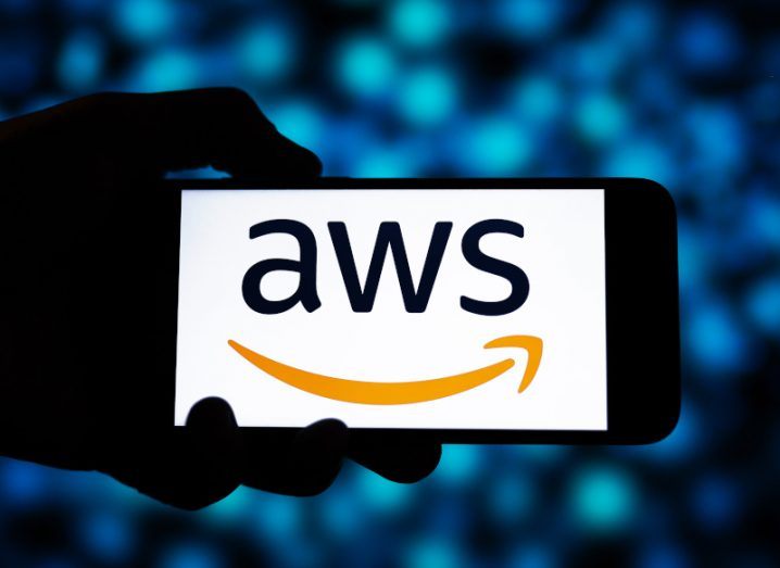 The Amazon Web Services logo on a smartphone screen, which is held in a person's hand with blue lights visible in the background.