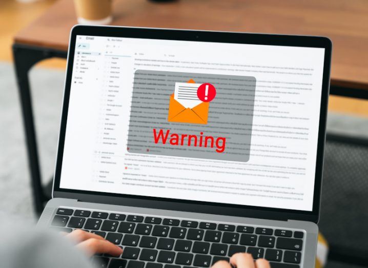 A person typing on a laptop with emails on the screen. There is a grey square in the centre of the screen with a warning symbol and an email icon on it.