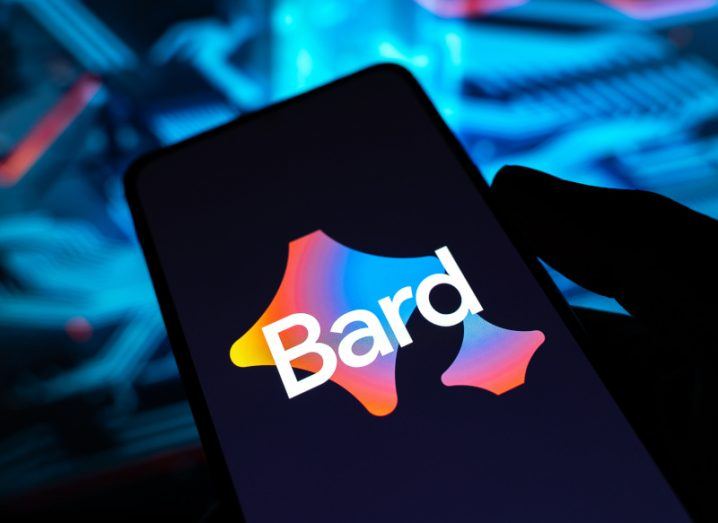 The Google Bard logo on a smartphone screen. The phone is in a person's hand in a dark room, with blue lighting in the background.