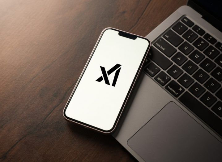 The xAI logo on a smartphone screen. The phone is laying on a grey laptop keyboard on top of a dark wooden surface.
