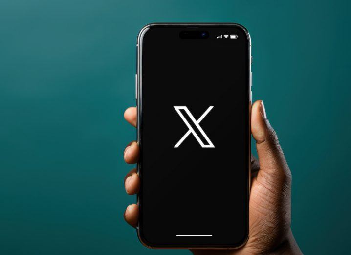 A person's hand holding a smartphone that has the X logo on its screen. There is a dark green background behind the phone.