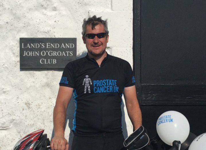 A man in a black t-shirt standing in behind a bike and in front of a wall. He is Duncan Bradley of Kyndryl.