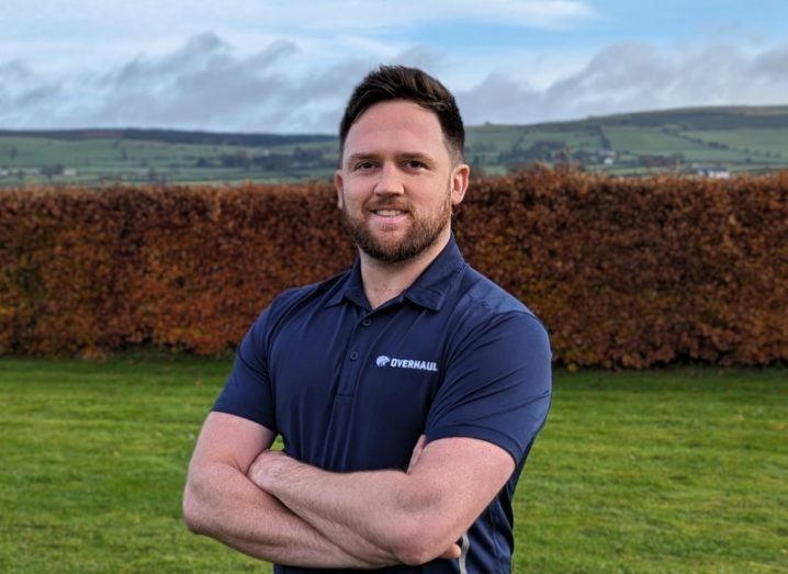 A man wearing a navy T-shirt with the word 'Overhaul' displayed on the front smiles at the camera with his arms folded. There is grass and a hedge behind him.