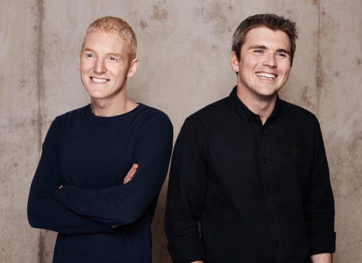 Patrick and John Collison stand next to each other, both wearing black and smiling, against a pale studio backdrop.