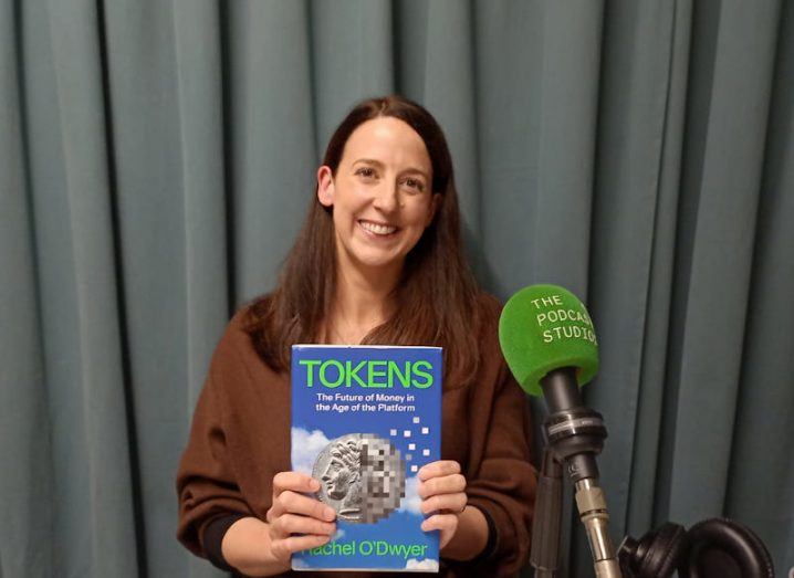 A woman sits in a desk with a microphone in front of her. She is holding a book titled Tokens.