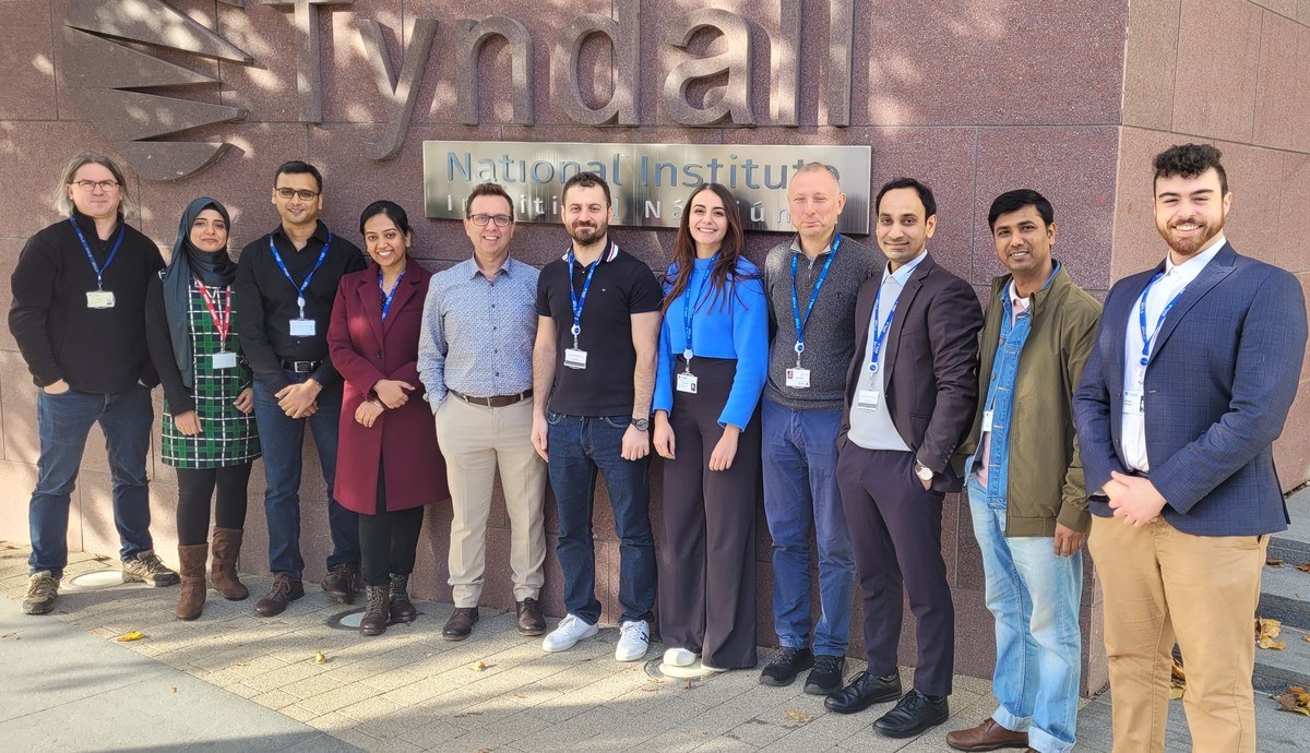 A group of men and women standing in front of a brown building with the Tyndall National Institute sign on the building wall.