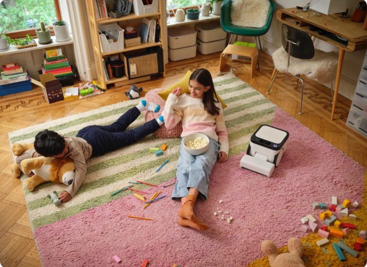 Two children lying on the floor of a room with the Matic robot cleaning around.