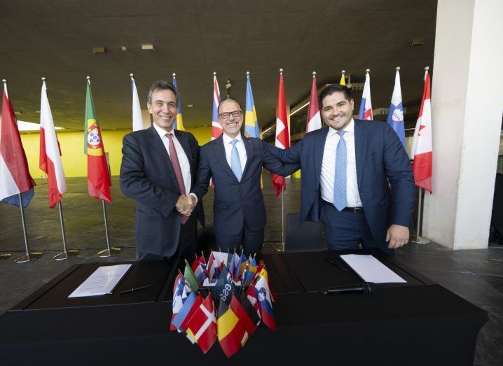 Three men wearing suits stand behind a table after seemingly signing a document. The flags of different countries are around them.
