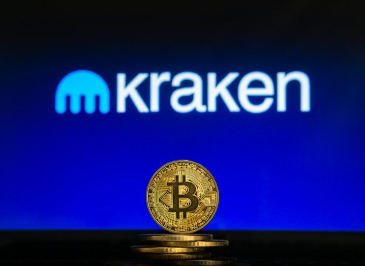 A physical bitcoin coin in gold with a background that has the Kraken logo on it.