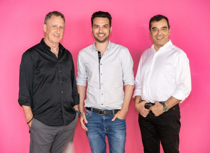 Three men stand next to each other and pose for a photo graph with hands in pockets in front of a pink wall.