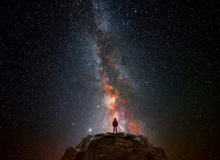 Silhouette of a person on top of a mountain staring at the vast expanse of the universe visible in a clear night sky.