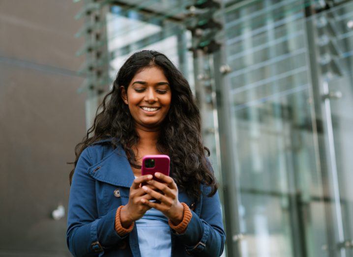 A young woman holds a smartphone in her hand and smiles at it.