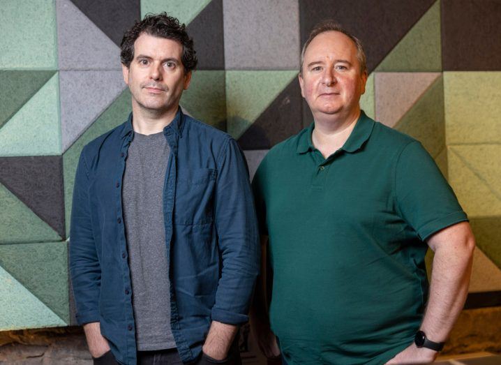 Two men stand next to each other in front of a wall with large triangular patterns.