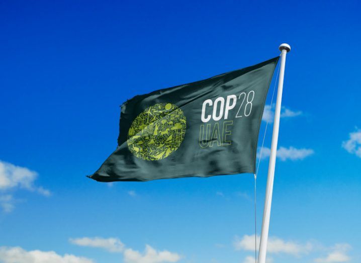 A green flag with the logo and words COP28 Dubai fluttering on a flag pole with a bright blue sky and scattering of clouds behind it.