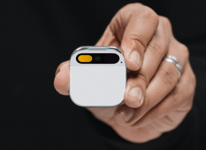 A small square device held in a person's hand. The device is the Humane AI Pin.