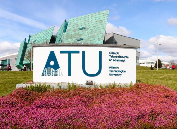 A sign for ATU, or Atlantic Technological University. The white sign is behind a pile of pink flowers, with a field of grass and a blue building behind the sign.