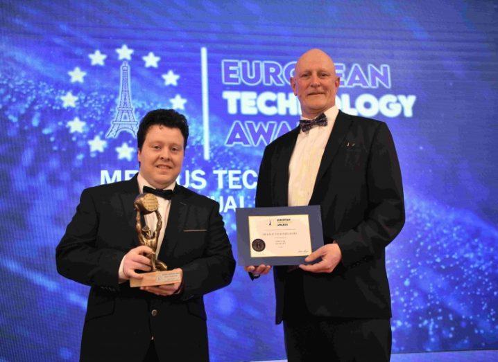 Two mean wearing black-tie suits stand on stage with their European Technology Award in their hands.