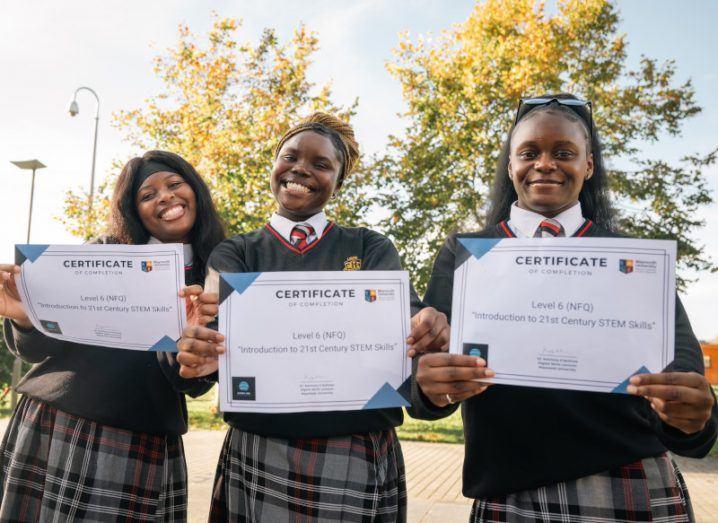 Three young women in school uniforms standing together and holding graduation certificates, with tress in the background.