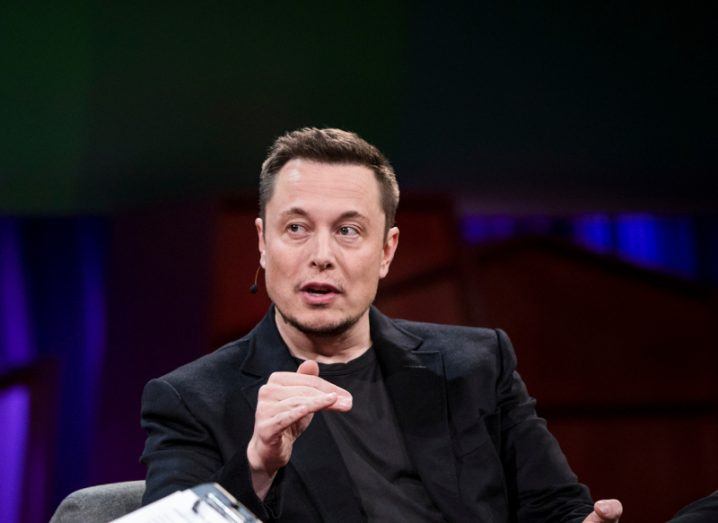 Elon Musk sitting on a chair and wearing a black suit, while speaking on stage.
