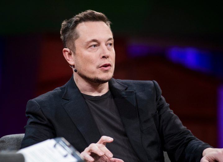 Elon Musk in a black suit sitting on a chair and speaking.