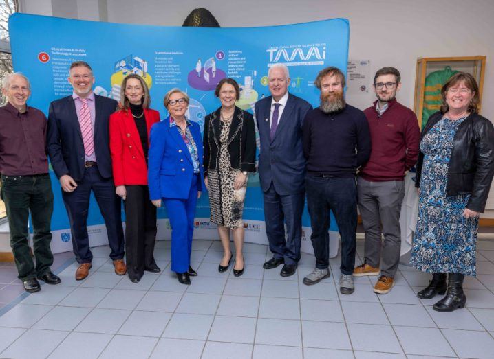 A group of men and women standing in a row in a room, in front of a blue banner that has the TMAI logo on it.