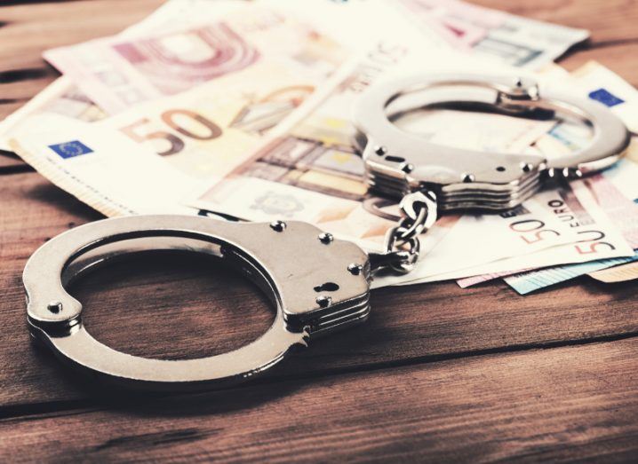 A pair of handcuffs on a wooden table. The handcuffs are on top of multiple euro notes. Used to represent the concept of financial crime.