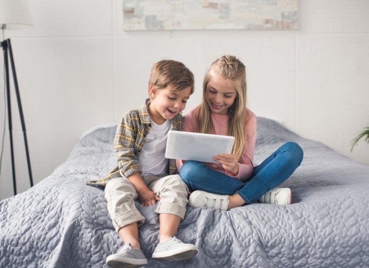 A boy and girl sitting on a bed and looking at a white tablet device.