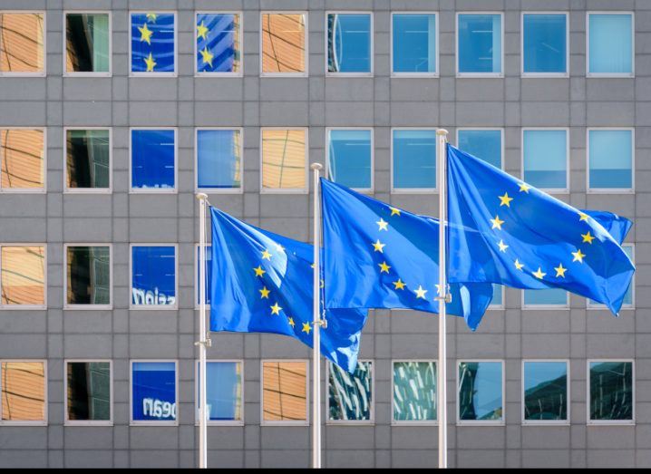 Three European Commission flags outside a building with a lot of windows.