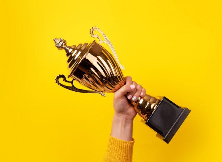 A hand holding up a gold trophy on a yellow background.
