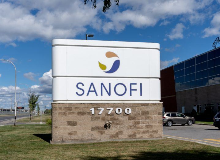 The Sanofi logo on a white sign in front of a building and a car, with a blue sky and clouds above.