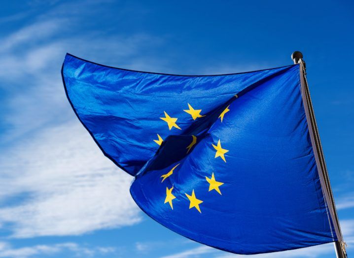 An EU flag waving on a flag pole, with a blue sky and some clouds in the background.