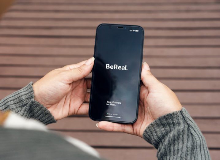 The BeReal app logo on the front of a smartphone screen. The phone is held in a person's hands and there is a wooden floor visible below.