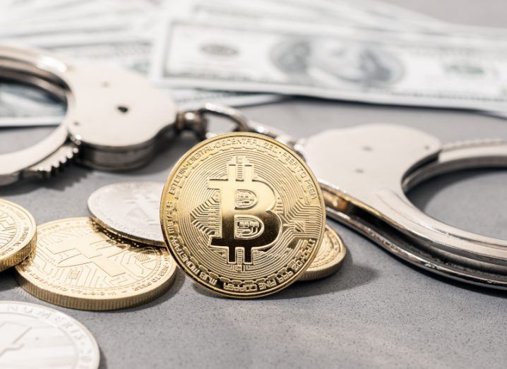 A gold coin with the bitcoin logo on it. The coin is on a table with coins, dollar notes and a pair of handcuffs behind it. Used to represent the concept of crime in the cryptocurrency sector.