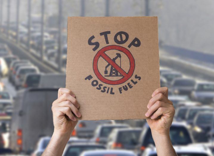 A cardboard sign that says 'stop fossil fuels' on it, held in a person's hands, with rows of vehicle traffic in the background.