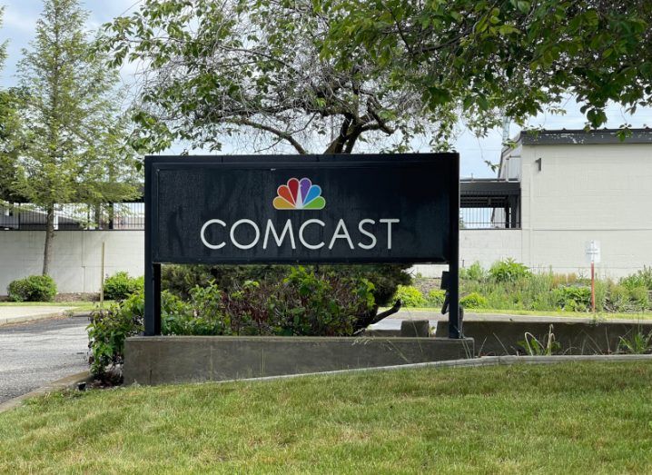 The Comcast logo on a black signpost, which is on a small piece of stone by some grass with a road next to it.