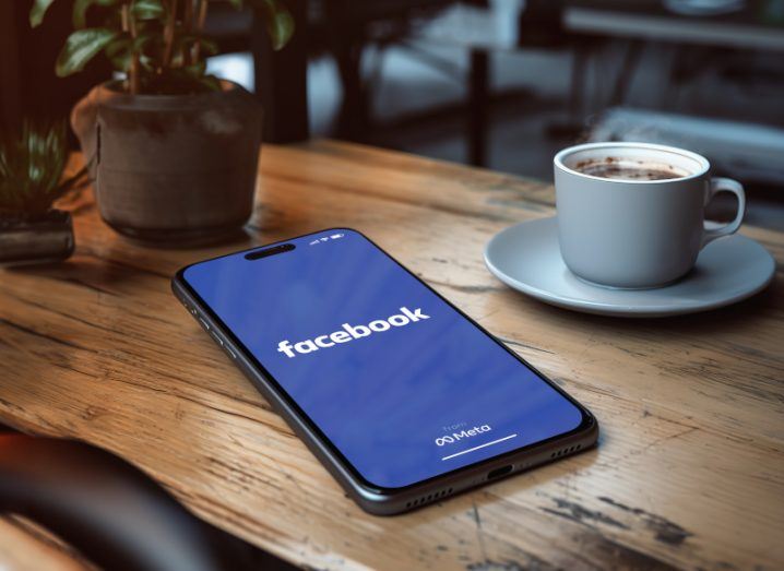 A smartphone laying on a wooden table next to a coffee cup and a plant. The Facebook logo is on the screen of the smartphone.