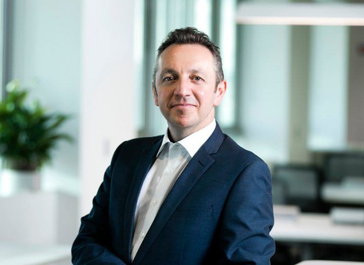 A man wearing a dark navy suit stands in an office setting smiling while holding his hands in front of him. He is Steve Coakley of BT Ireland.