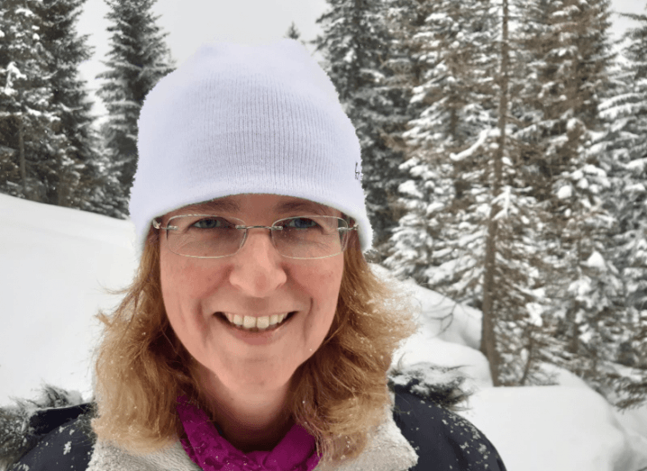 Prof Louise Harra wears a white woolly hat and puffy coat with snow and mountain and trees behind her.