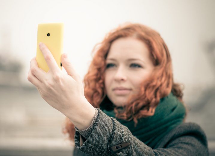 Woman holds a smartphone in front of her face as if to take a selfie. She is in the outdoors and the phone is in focus.