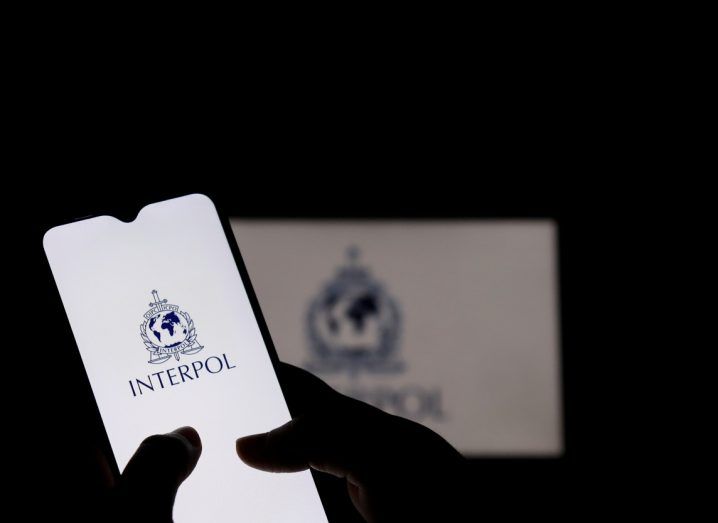 Person holding a phone with the Interpol logo on the screen. The setting is a dark room and the logo can also been seen on a wall in the background.