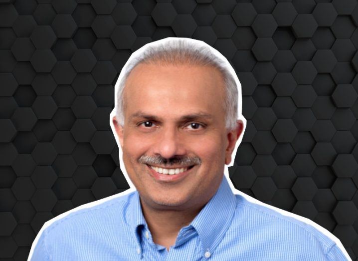 A man wearing a blue shirt smiles at the camera in front of a black background made up of hexagonal shapes. He is Apurva Mehta, CTO of Versa Networks.