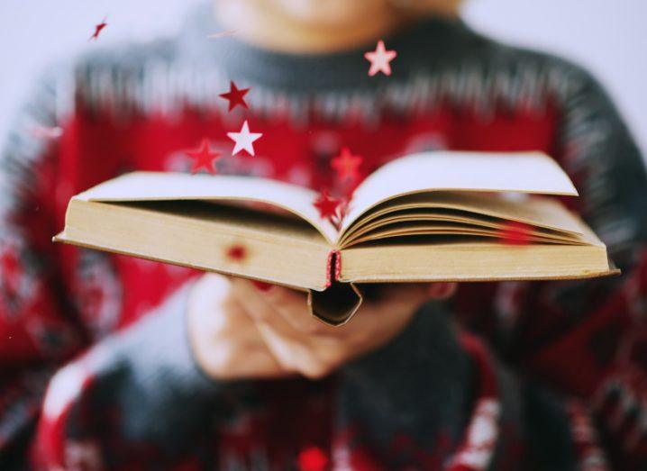 A close-up of an open book with red stars flying around it. It is held out by a woman in a Christmas jumper who is blurry.