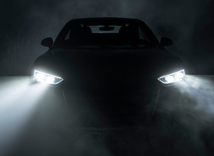 A dark image of a black car in a dark garage with the headlights on.