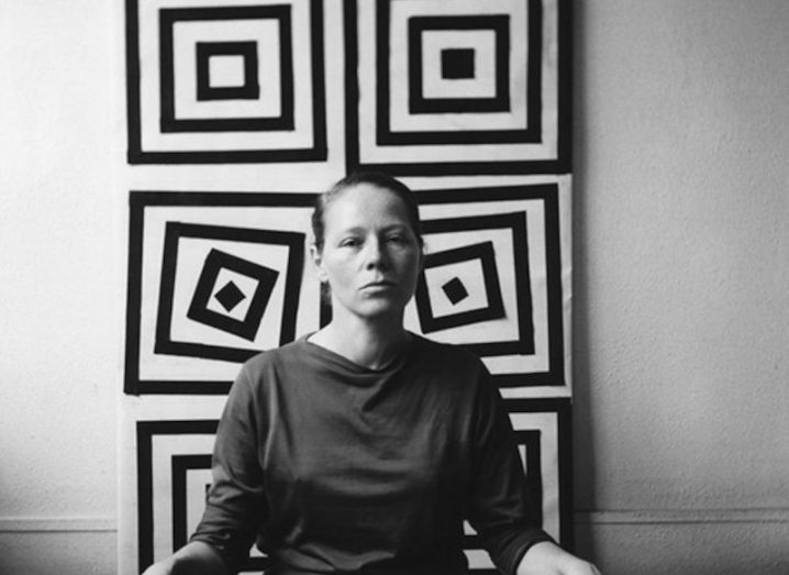 A black and white image of a woman sitting in front of a wall that has six artistic images of squares behind her. She is Vera Molnár.