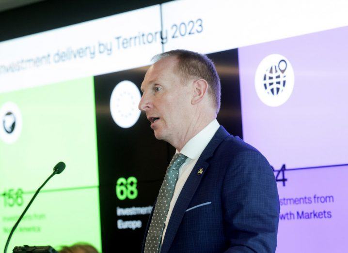 A man wearing a suit standing on a stage and speaking into a microphone, with graphs on a wall behind him. He is Michael Lohan of IDA Ireland.