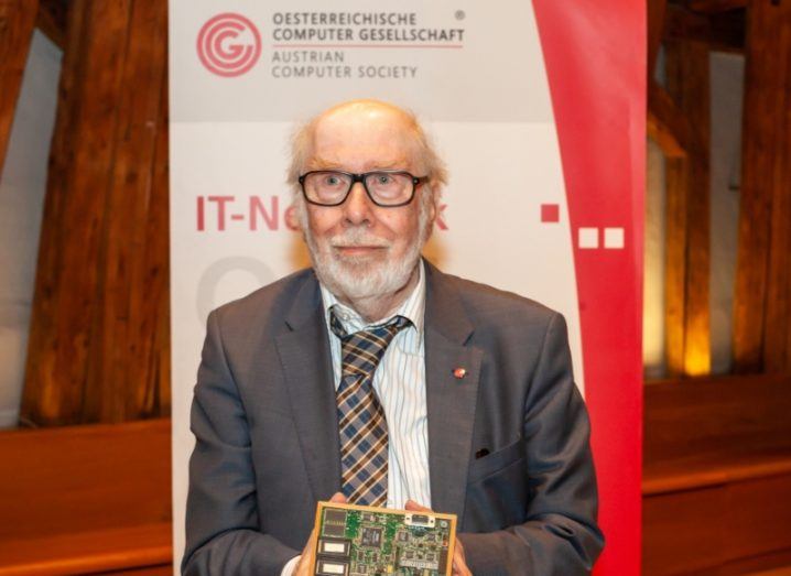 Niklaus Wirth in a grey suit, standing in front of a sign with the Austrian Computer Society logo on it. Wirth is holding a small electronic device in his hands.