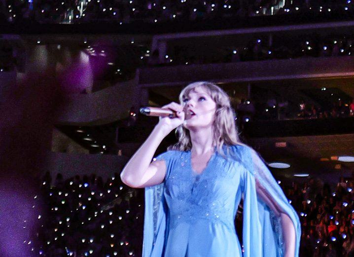 Taylor Swift wearing a blue dress and holding a microphone, singing on a stage with a crowd behind her.