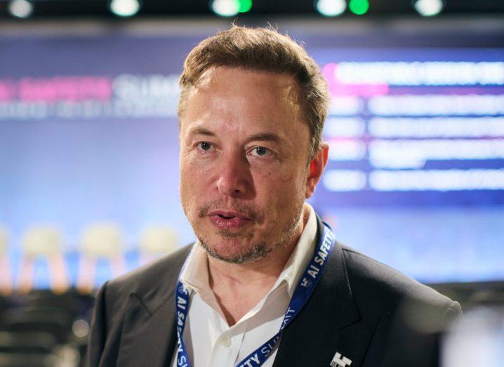 Elon Musk wearing a black suit with a large screen visible in the background.