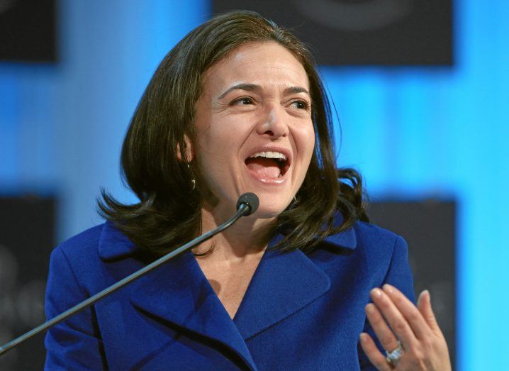 Photo of Sheryl Sandberg speaking on stage at an event. She appears to be smiling while talking.